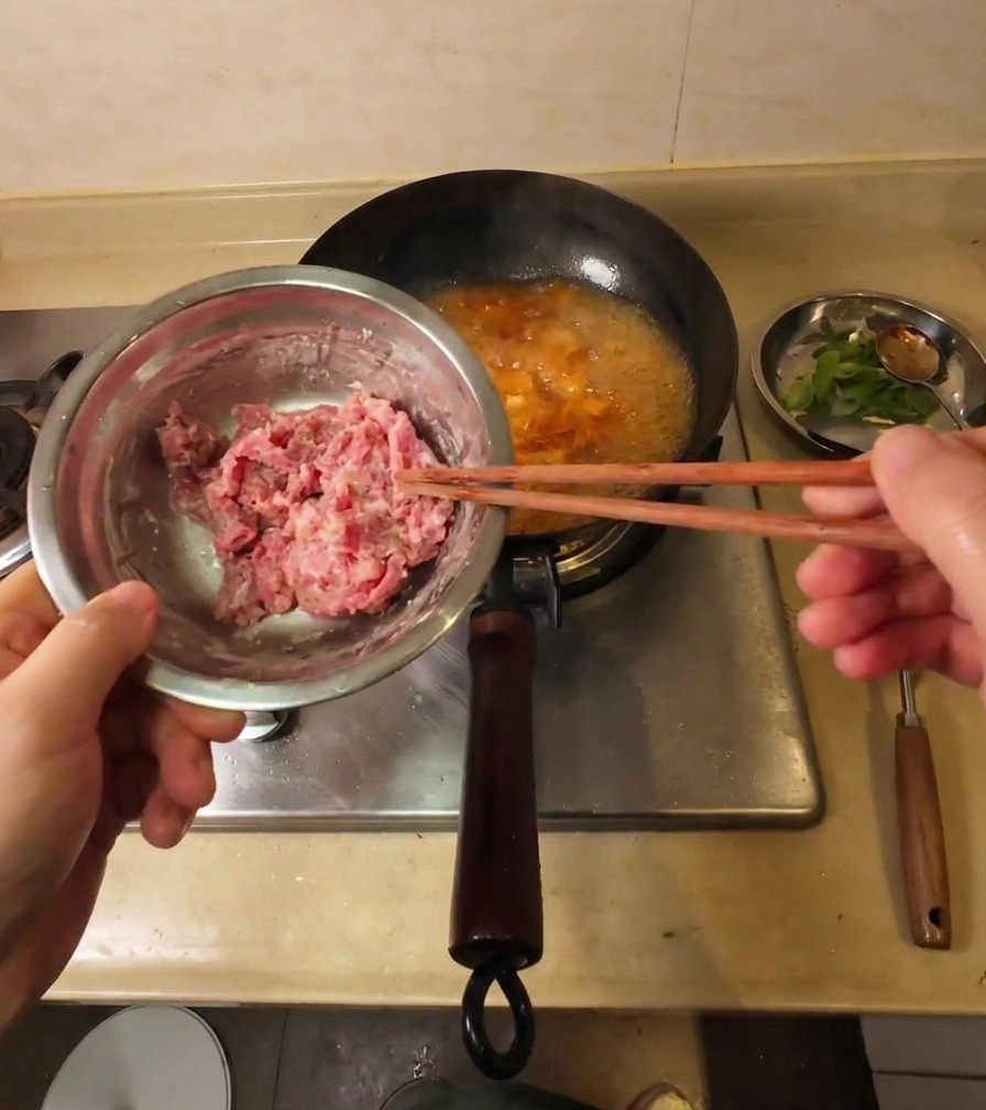 A hand using chopsticks to lift thin starch-coated raw beef slices from a metal bowl over a pot of bubbling soup.