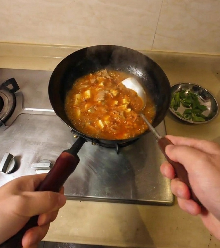 A person stirring a bubbling, thick orange-red soup containing tofu and beef pieces in a black wok on a stove.