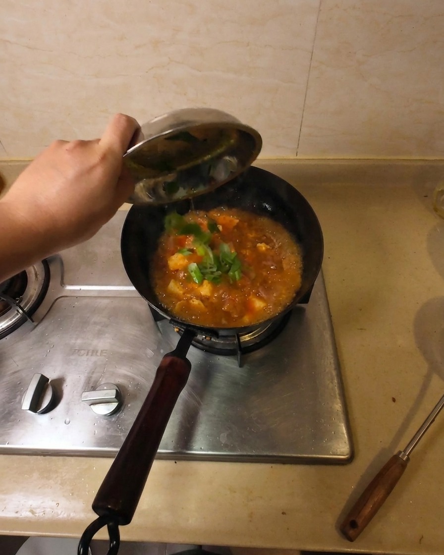A hand pouring fresh chopped green onions from a small metal bowl into a wok of simmering beef and tofu soup.