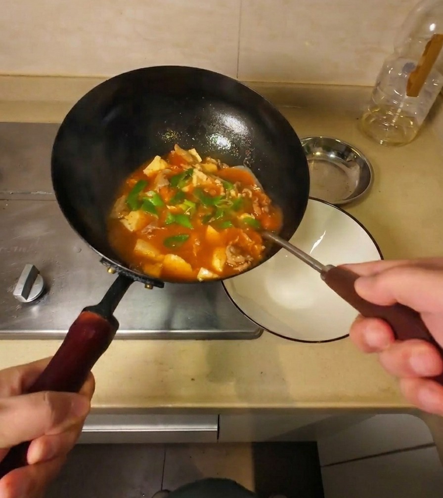 Finished thick beef and tofu soup being poured from a tilted black wok into a white ceramic serving bowl.