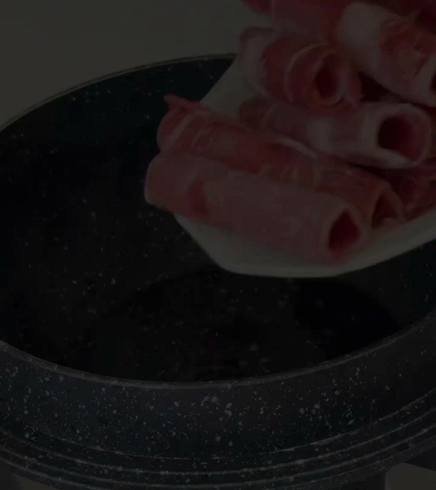 A hand placing thin, rolled slices of raw red beef brisket into a speckled black non-stick pot.