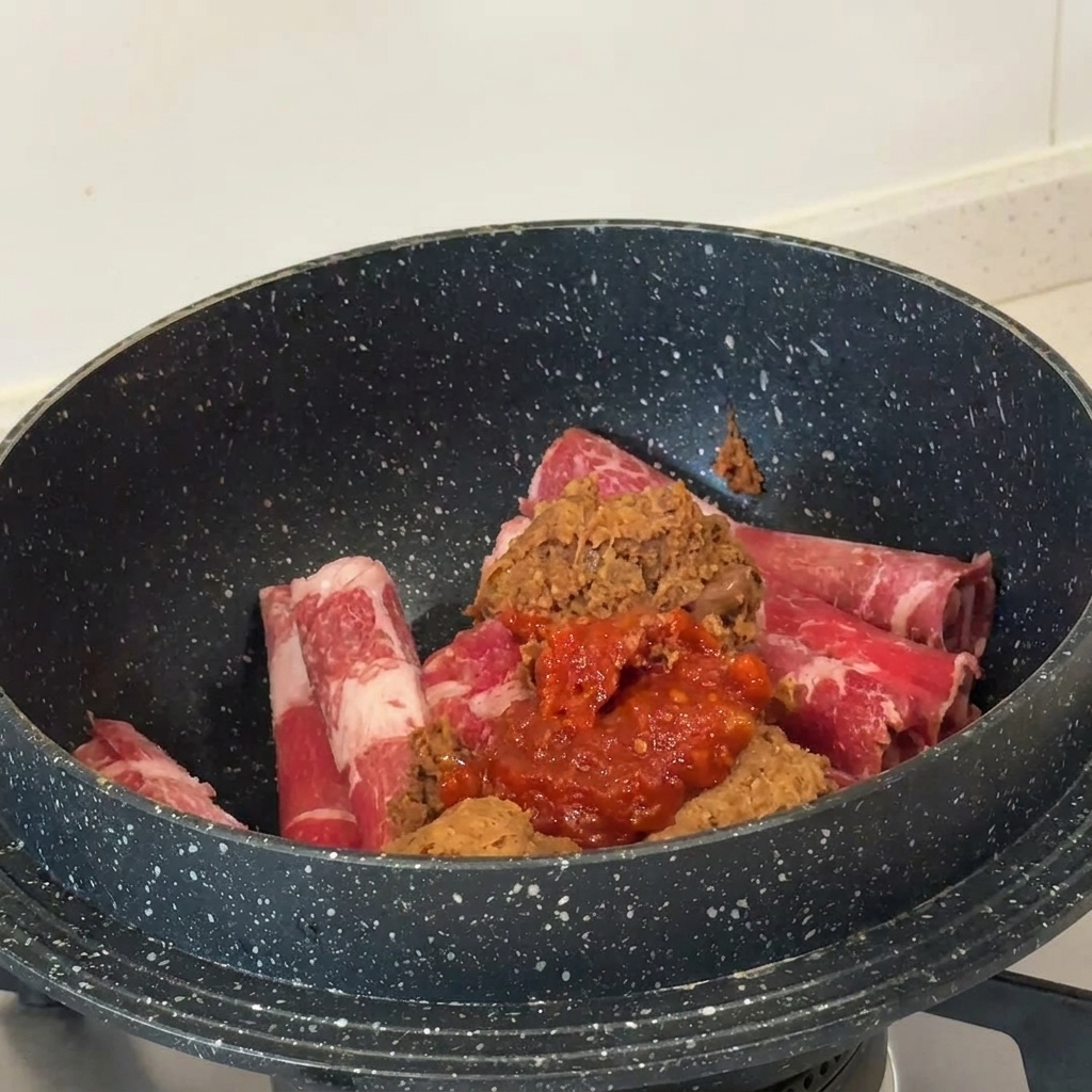 Mounds of brown soybean paste and vibrant red chili paste sitting on top of raw beef slices in a dark pan.