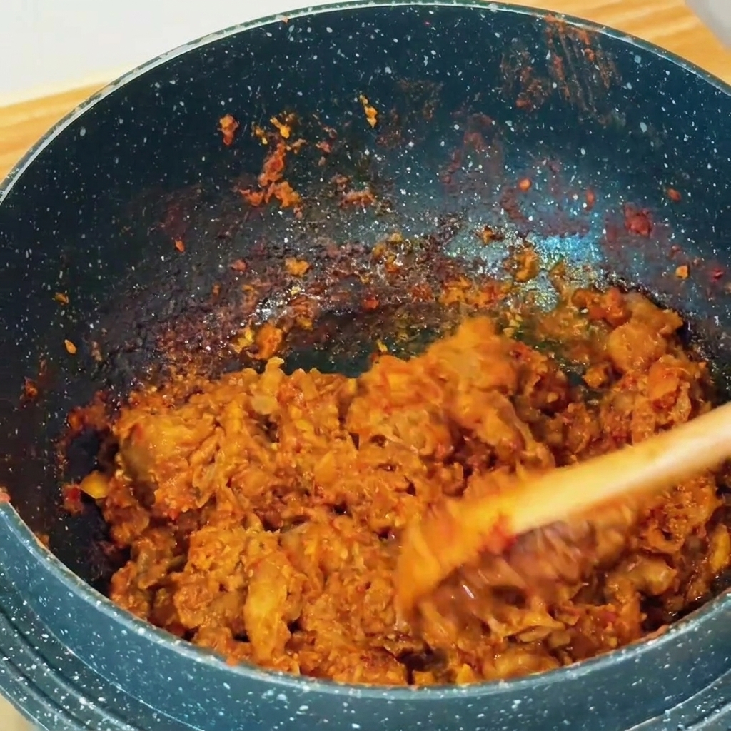 Thinly sliced beef being stir-fried with reddish-brown soybean paste in a dark speckled pan using a wooden spatula.