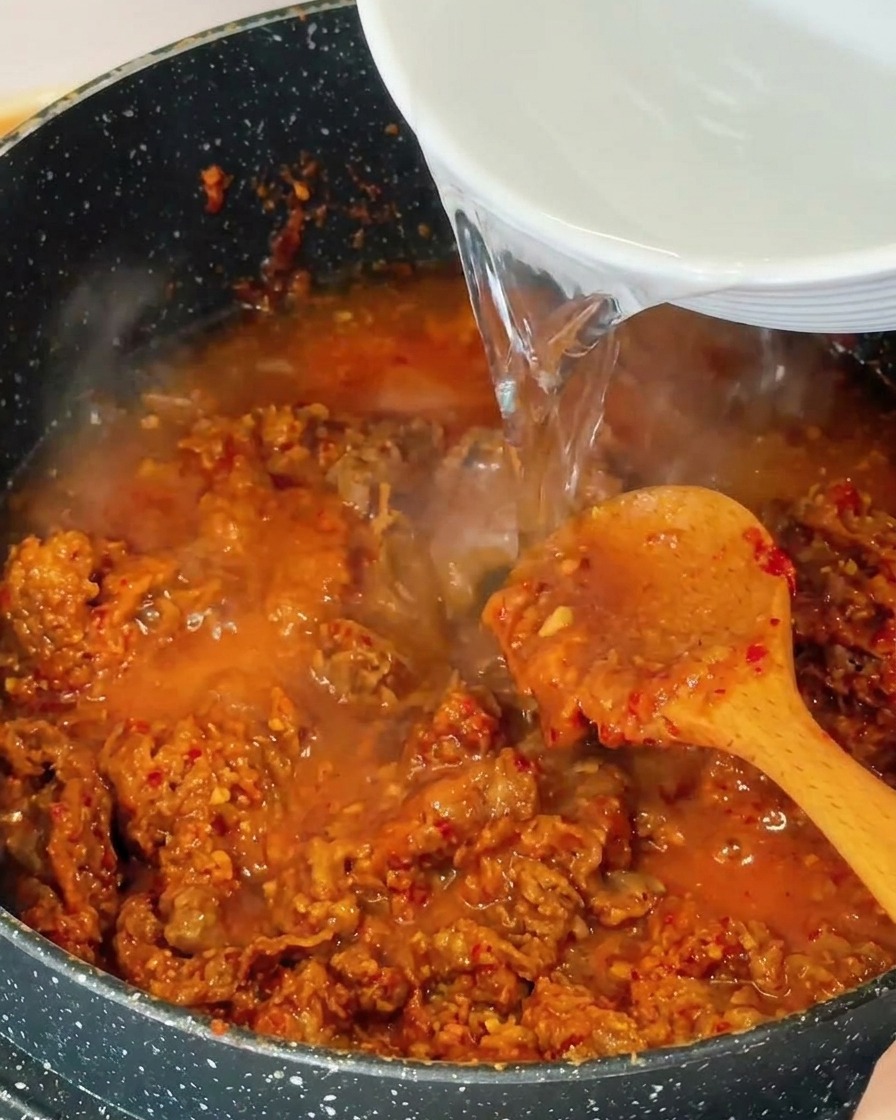 Clear water being poured from a white bowl into a pan containing stir-fried beef and bubbling red-brown broth.