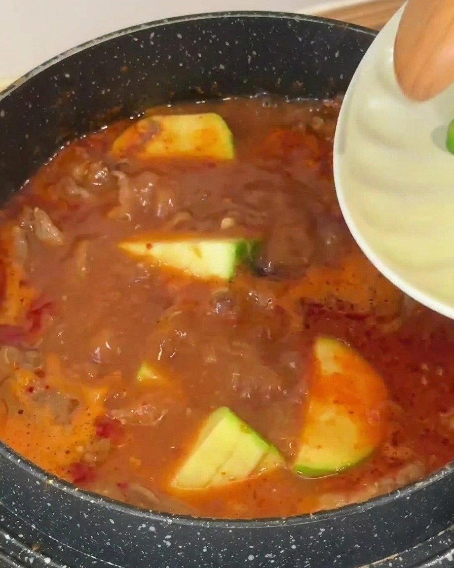Several slices of green-skinned zucchini being dropped into a bubbling, reddish-brown soup base.