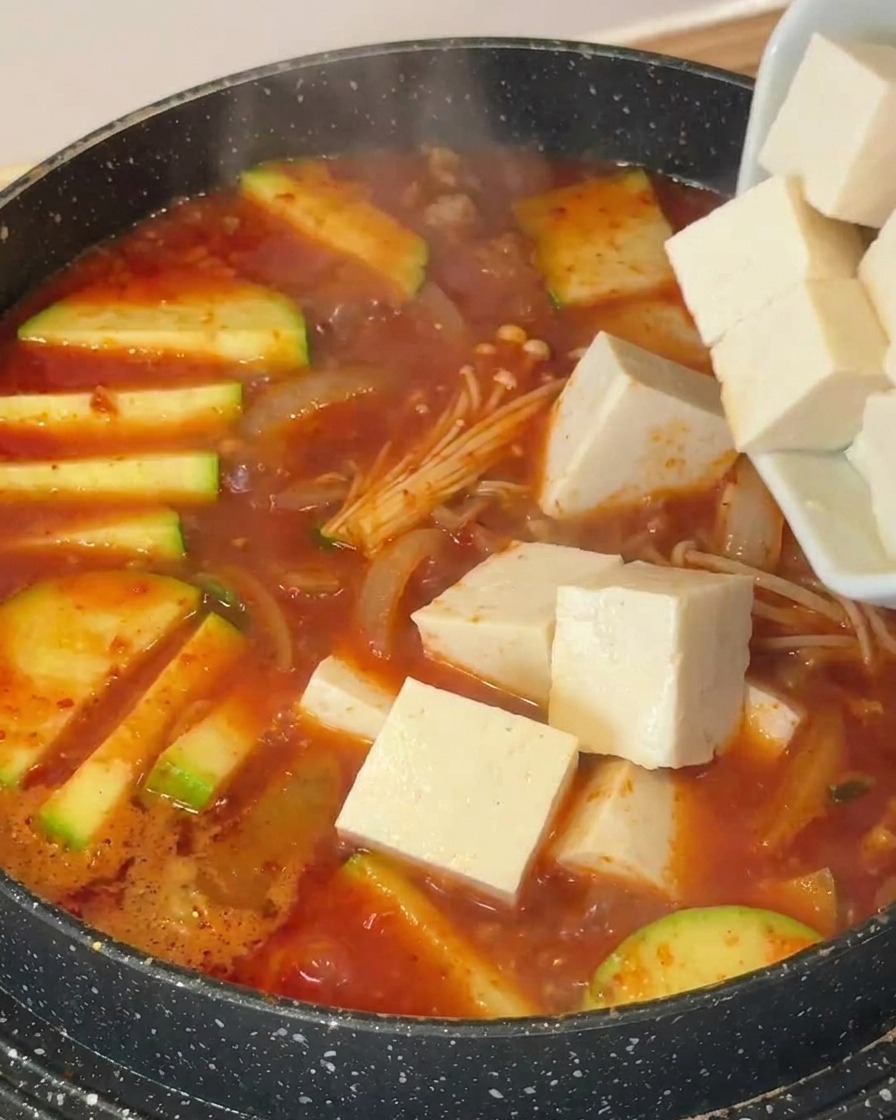 Cubes of white tofu being added to a pot filled with bubbling soup, zucchini, and enoki mushrooms.