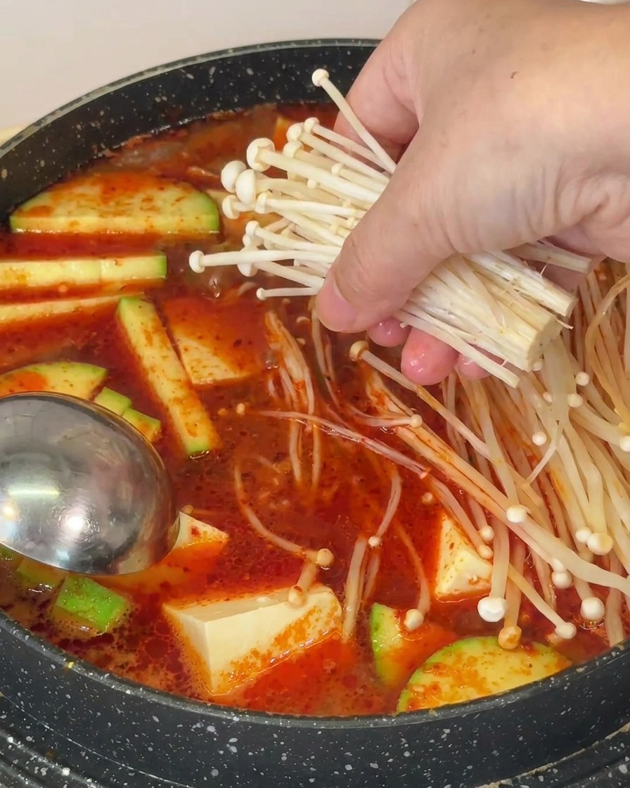 A hand placing a bundle of long, thin enoki mushrooms into a boiling pot of red soup containing tofu and zucchini.