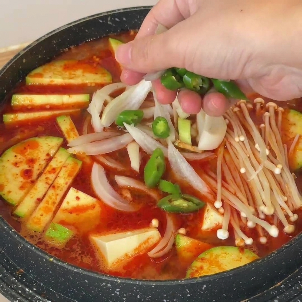 Hand sprinkling green chili slices over a pot of Korean-style soybean paste soup filled with mushrooms, tofu, and zucchini.
