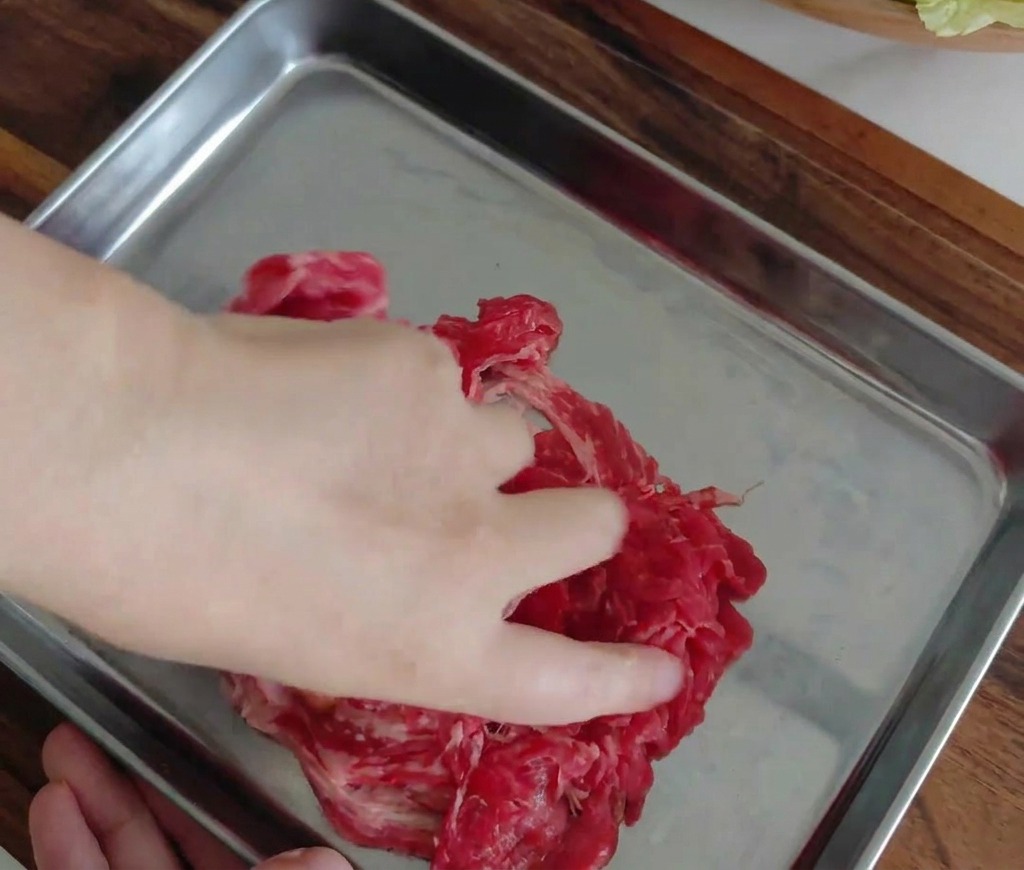 A hand mixing the marinated beef slices in a metal tray.