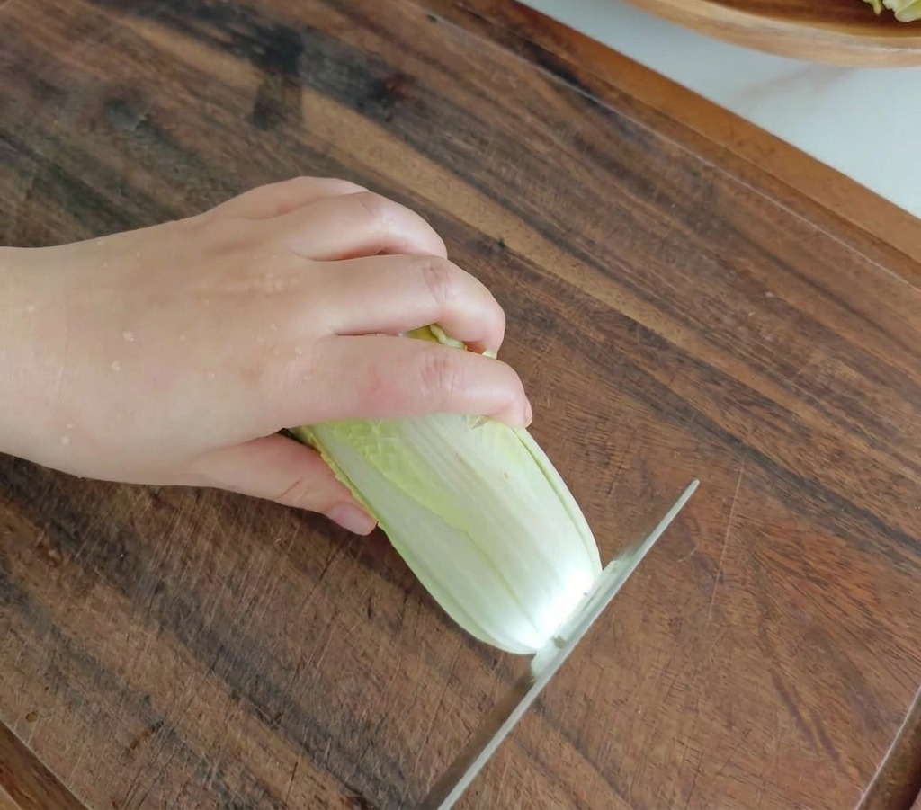 A kitchen knife slicing through a fresh head of baby cabbage on a dark wooden cutting board.