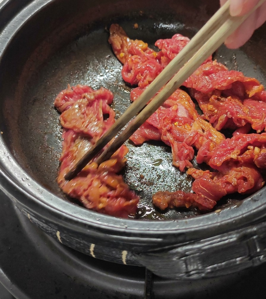 Sliced beef being stir fried in a dark pot with wooden chopsticks.
