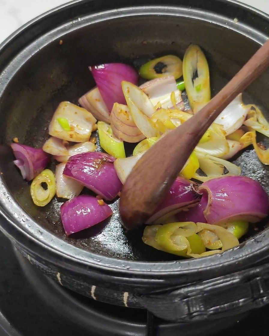Wooden spoon stirring large pieces of red onion and green onion whites in a dark stone pot.