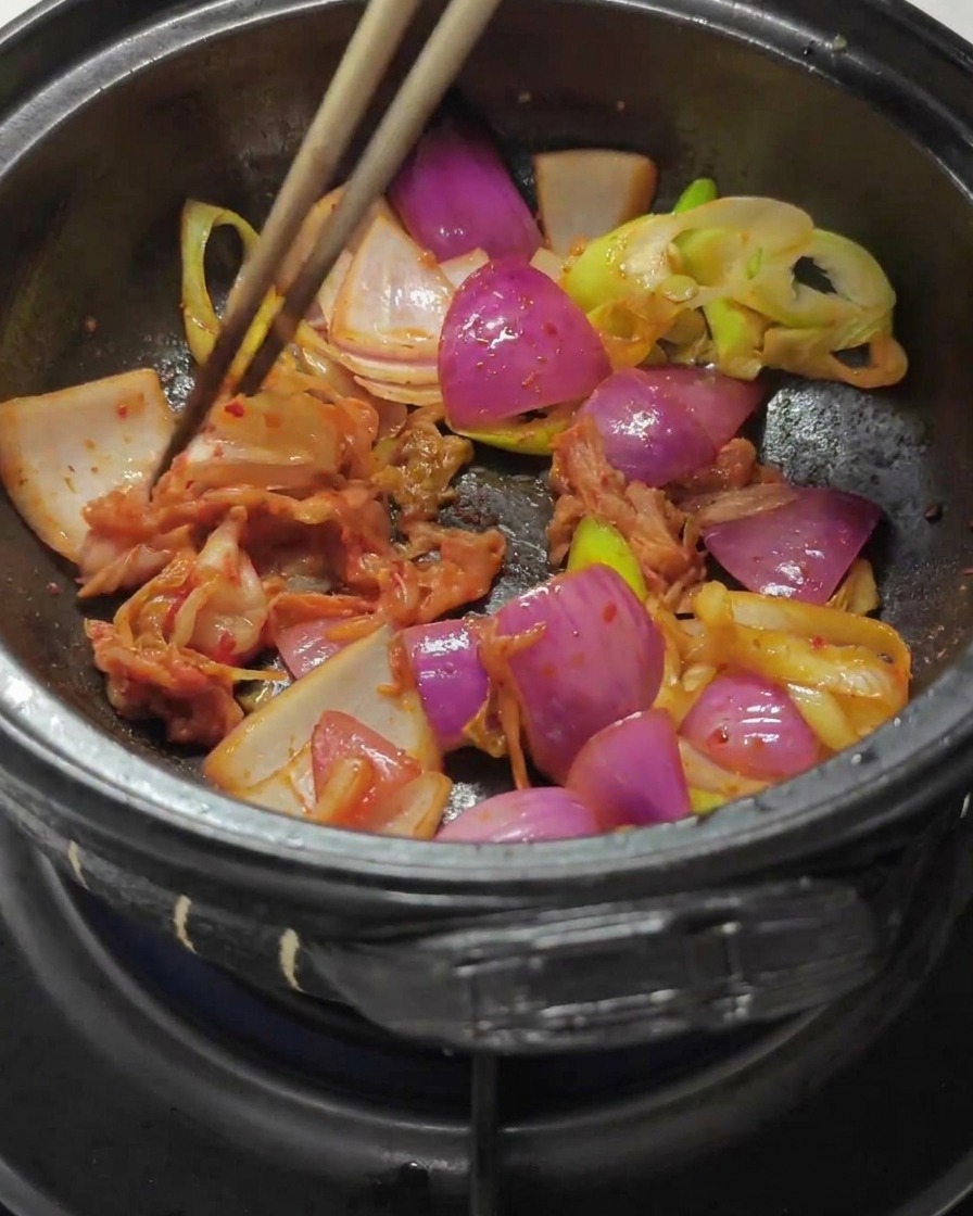 Chopsticks mixing chopped cabbage kimchi and red chili paste into the sautéed onions in a stone pot.