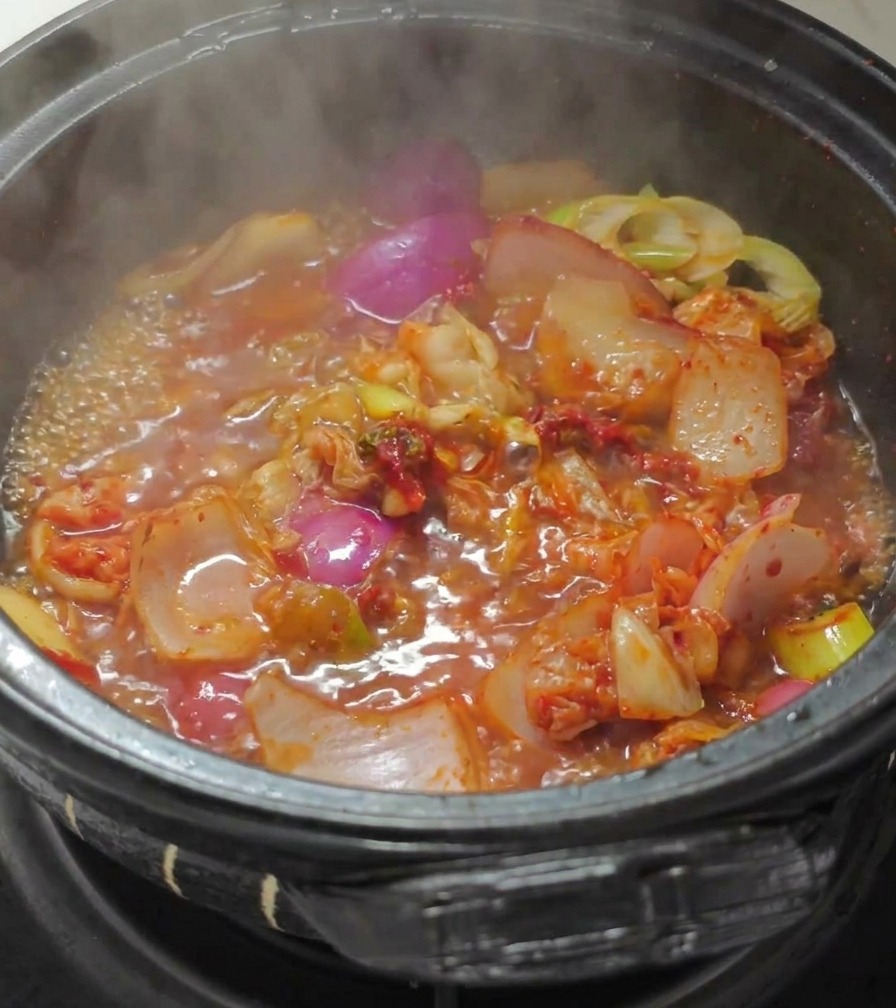 Boiling water being poured into a black stone pot containing sautéed kimchi, red onions, and green onions in a vibrant red broth.