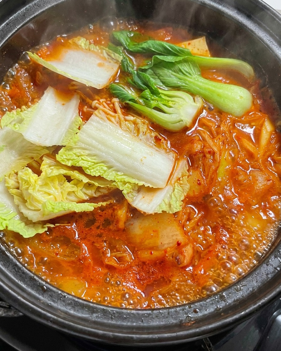 Fresh cabbage leaves being added to a bubbling red stew in a stone pot.