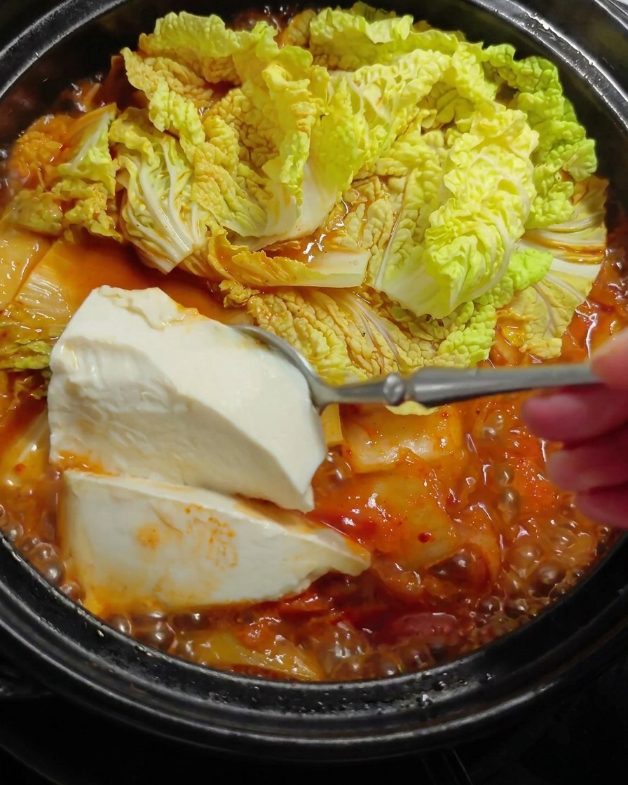 A large spoonful of soft white tofu being gently lowered into a hot, bubbling red stew with cabbage leaves.