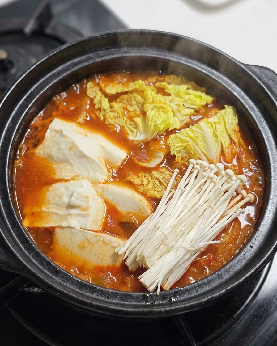Sliced beef, enoki mushrooms, and green onions being added to the top of a bubbling red stew.