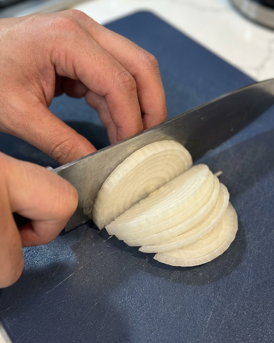 A close-up shot of hands using a sharp chefs knife to slice a halved white onion into thin strips on a navy blue cutting board.
