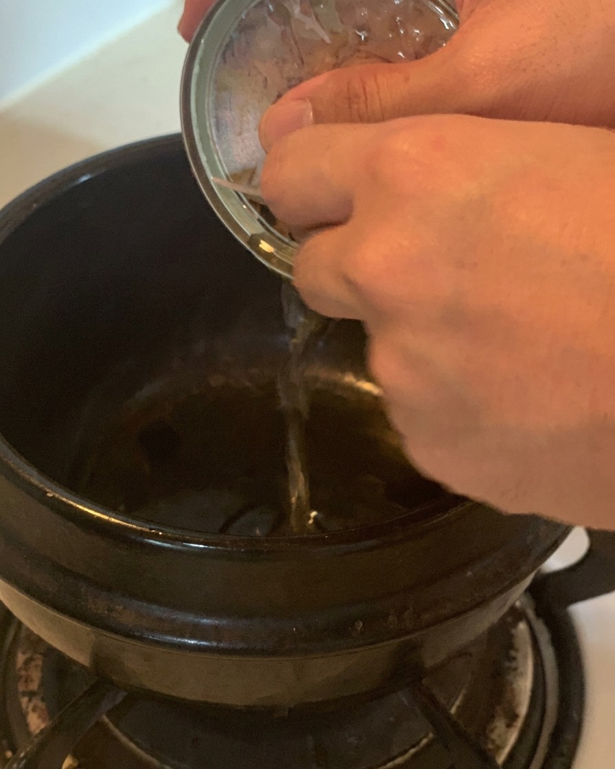 A hand pouring clear oil from a silver tuna can into a black ceramic cooking pot on a stovetop.