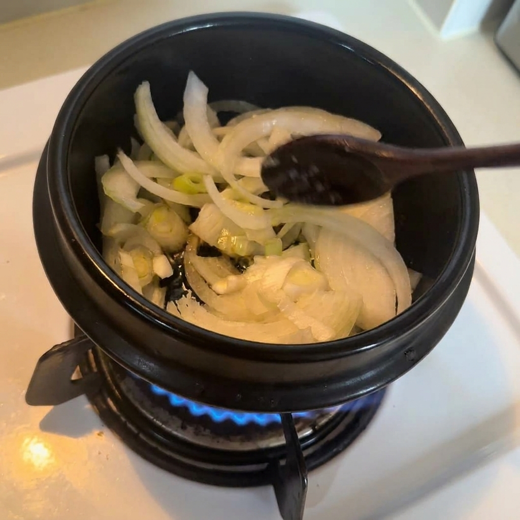 Sliced onions and scallions being stirred with a wooden spoon in a black stone pot as they sauté on the stove.