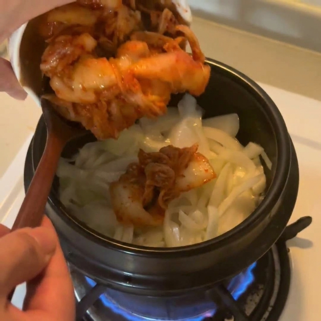 Chopped red cabbage kimchi being poured from a white ceramic bowl into a pot of sautéed onions.