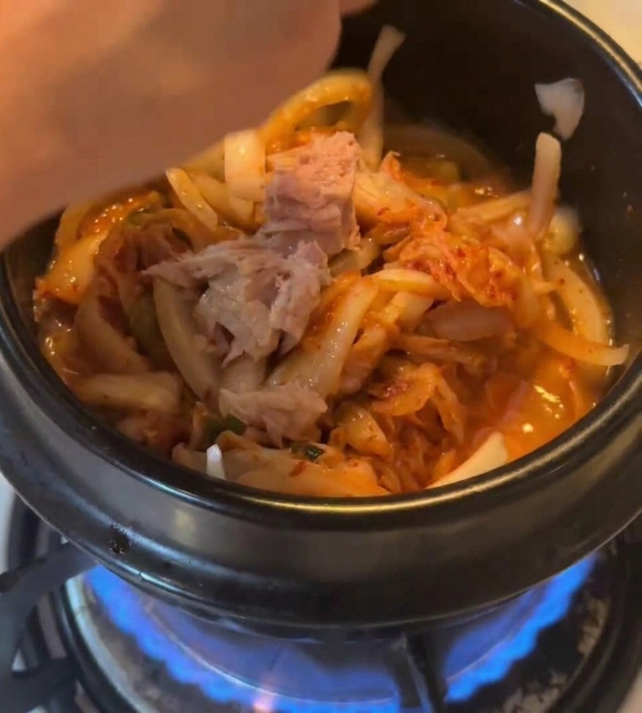 Chunks of canned tuna being added into a black stone pot containing stir-fried kimchi and onions over a blue gas flame.