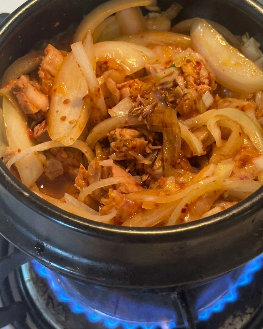 A close-up of dark soy sauce being drizzled over tuna, kimchi, and onions in a cooking pot.