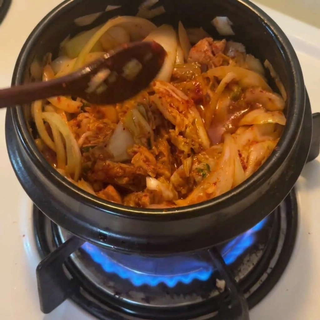 A wooden spoon sprinkling red Korean chili powder over the tuna and kimchi mixture in a black pot.