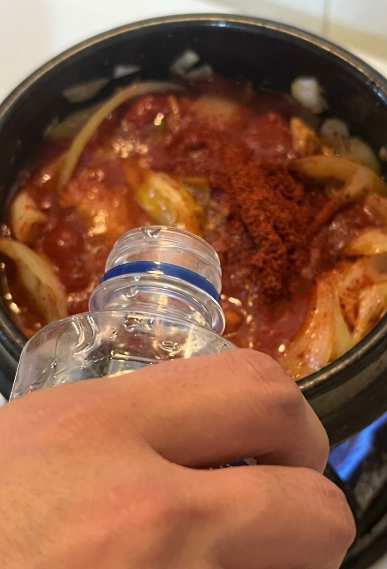 Water being poured from a clear bottle into a pot of red kimchi and tuna stew over a gas stove.