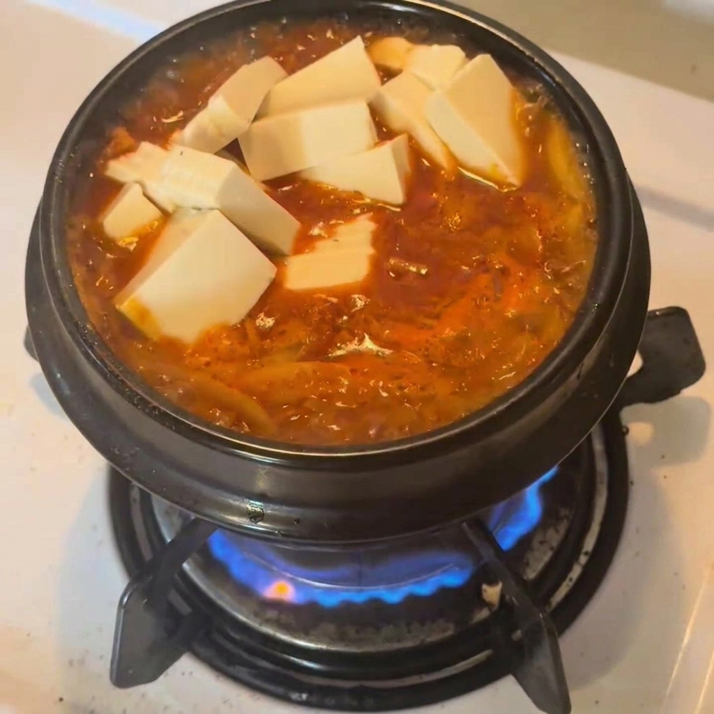 Cubes of fresh white tofu resting on top of a bubbling, bright red kimchi tuna stew in a black ceramic pot.