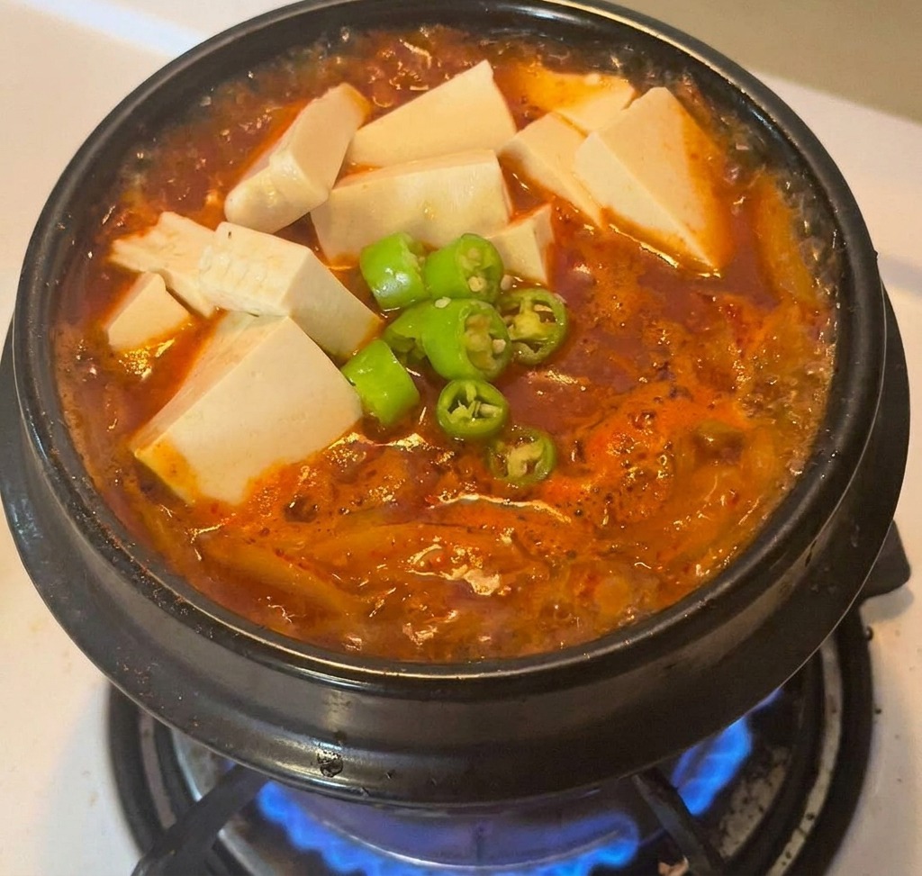 A black stone pot filled with vibrant red kimchi stew, topped with several large cubes of white tofu and a pile of sliced green chilies in the center.