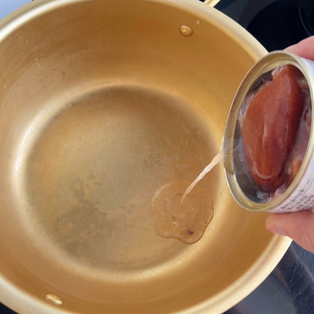 Oil being poured from a small tin of canned tuna into the center of a gold-colored cooking pot.