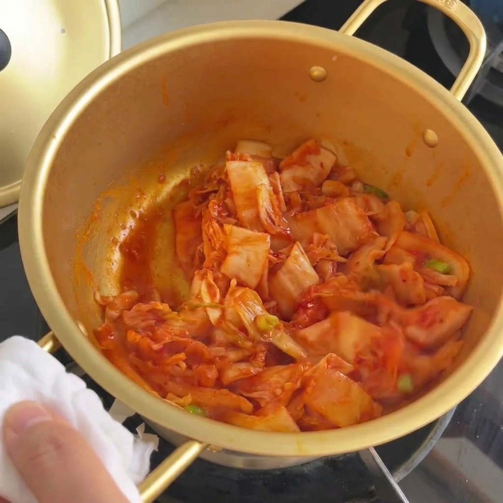 Stir-fried kimchi and green onions being mixed together in a gold pot until the cabbage looks translucent.