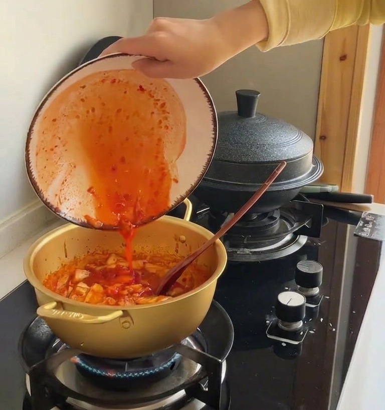 Vibrant red kimchi juice being poured from a ceramic bowl into a gold pot filled with stir-fried cabbage.