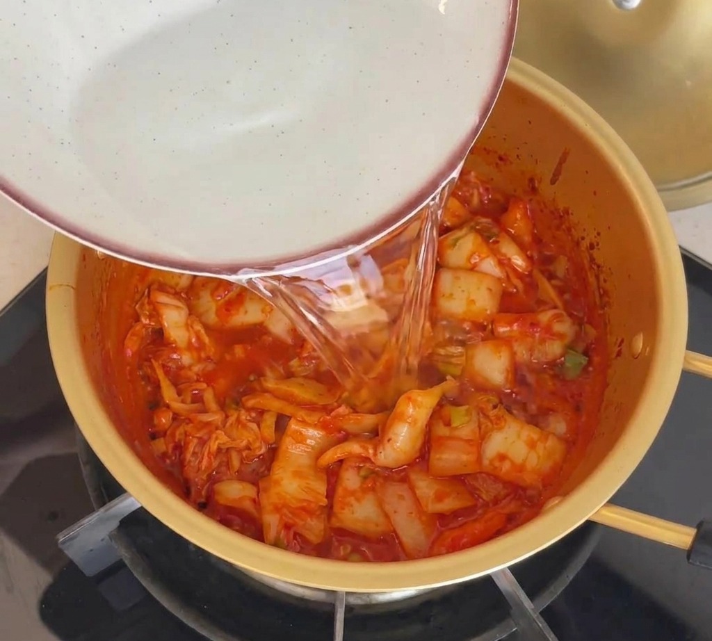 Water being poured from a ceramic bowl into a gold-colored pot containing red sautéed kimchi.