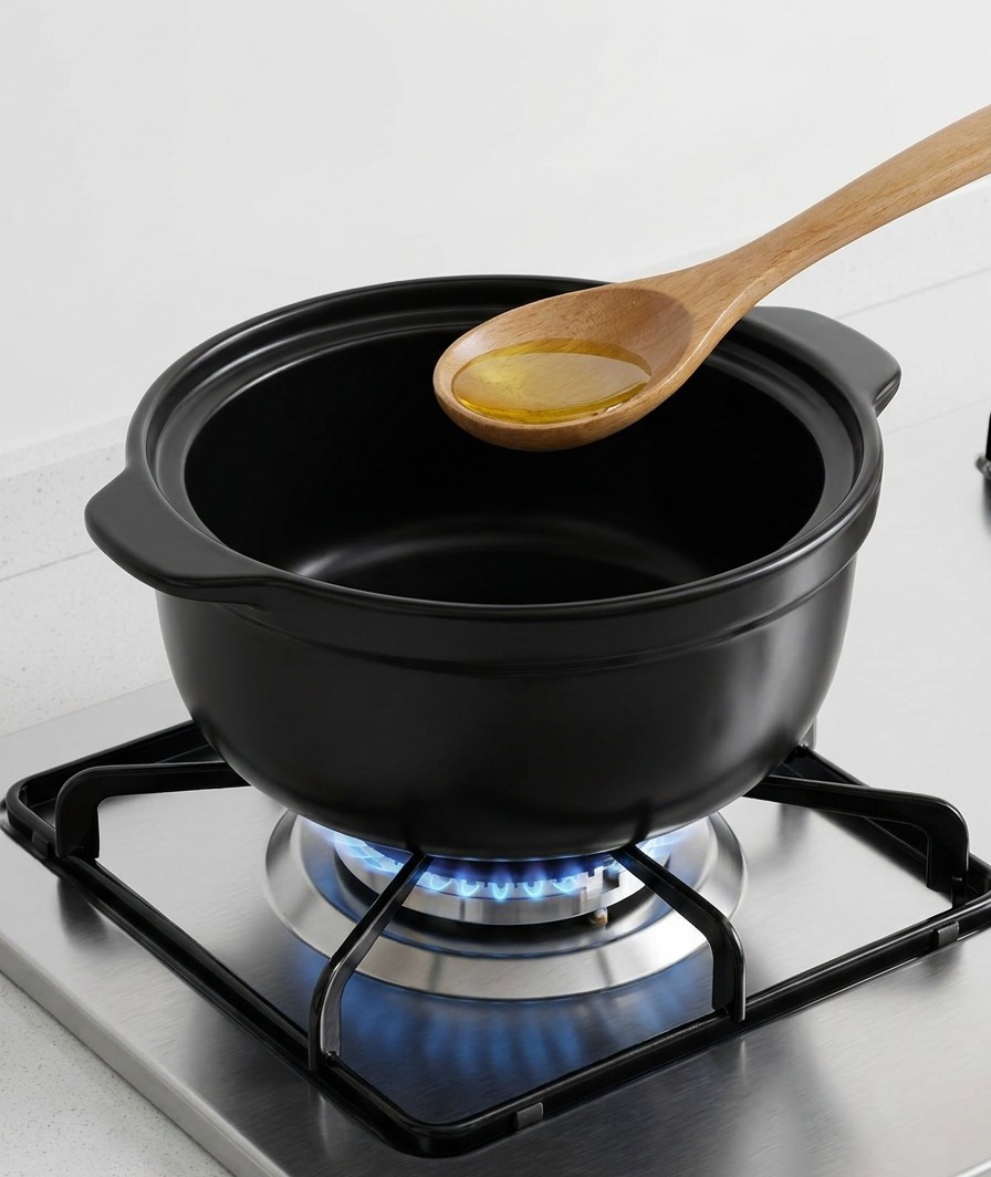 A wooden spoon pouring golden sesame oil into the bottom of a dark ceramic cooking pot over a gas flame.
