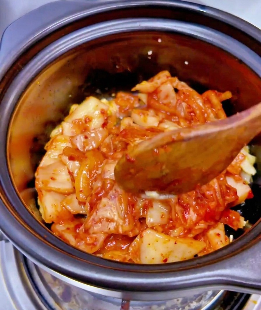The liquid from a can of tuna being poured onto a mixture of chopped kimchi and garlic in a black ceramic pot.