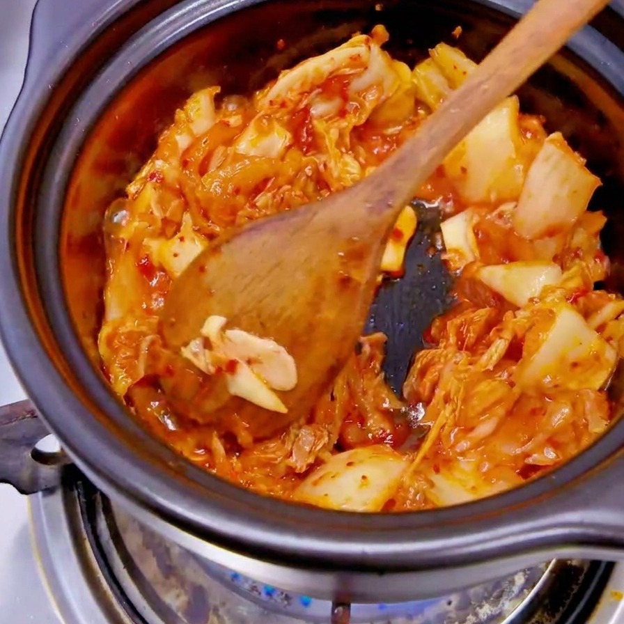 A hand wearing a black glove dropping white onion slices into a pot containing red kimchi.