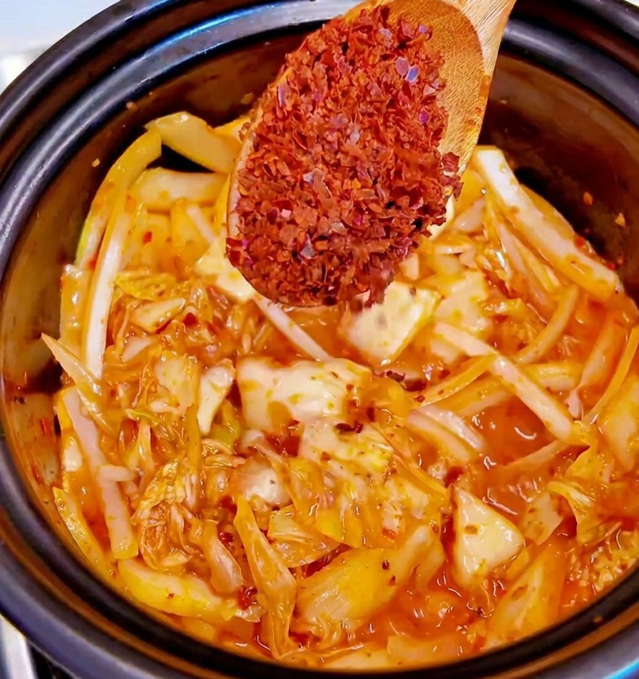 A wooden spoon holding coarse red chili flakes over a pot of stir-frying kimchi and onions.