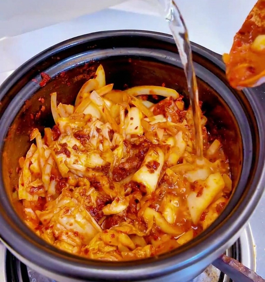 Water being poured from a container into a black pot filled with spicy kimchi base.