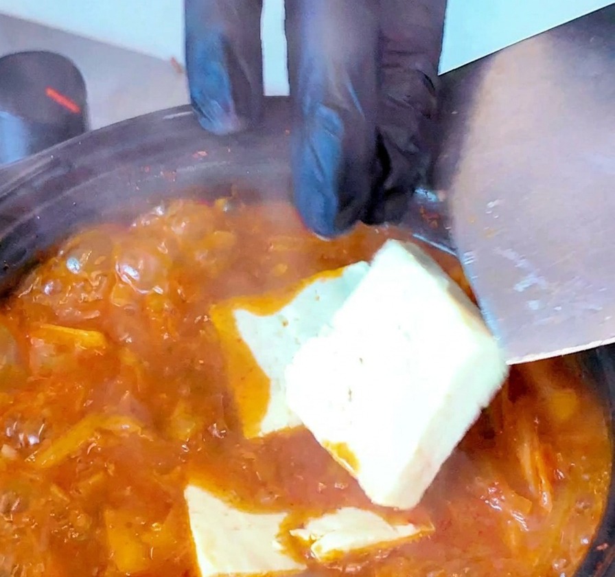 Slices of white tofu being lowered into a bubbling red kimchi soup using a spatula.