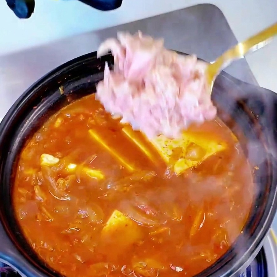 Canned tuna meat being added by the spoonful to a pot of boiling kimchi and tofu soup.