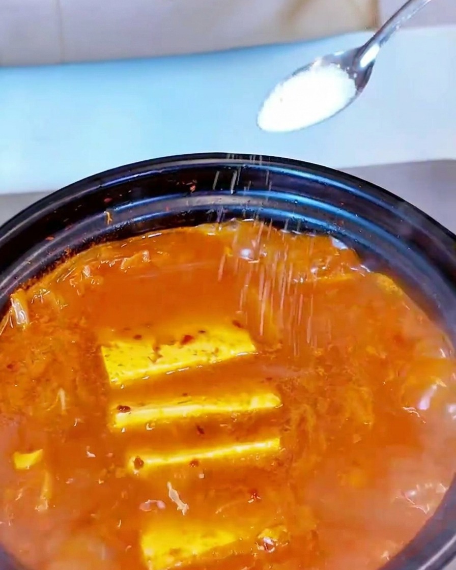 White salt being sprinkled from a small spoon onto the surface of a simmering red soup.