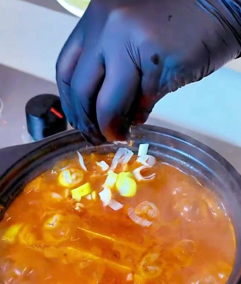 A hand in a black glove sprinkling a handful of chopped green onions into a steaming pot of red-orange kimchi soup.