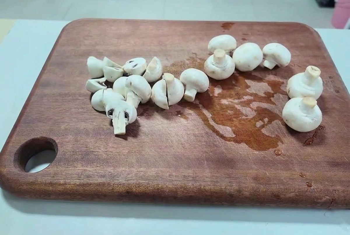 Fresh white button mushrooms being sliced into halves and thick pieces on a dark wooden cutting board.