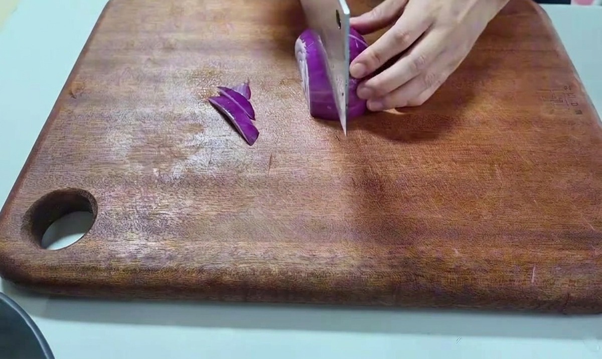 A hand holding a red onion and slicing it with a large cleaver on a wooden board.