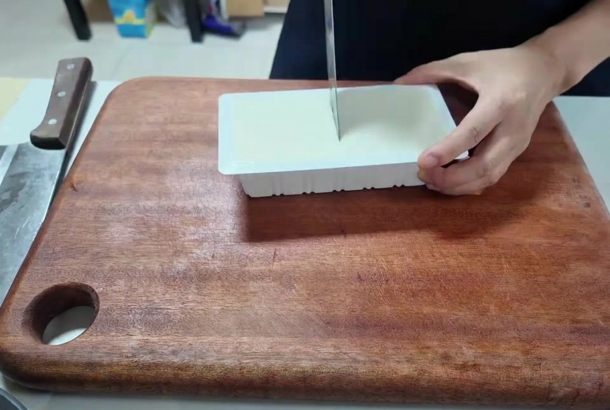 A person using a kitchen knife to slice a rectangular block of white tofu into small cubes on a wooden cutting board.