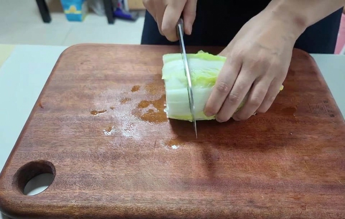 Close-up of hands using a chefs knife to chop a stack of fresh napa cabbage leaves on a dark wooden cutting board.