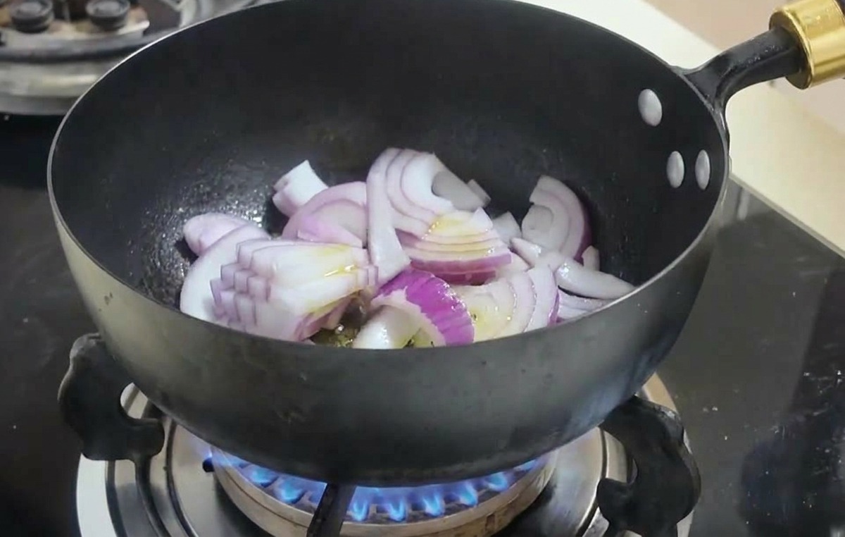 Sliced red onions being stir-fried in a black wok over a blue gas flame.