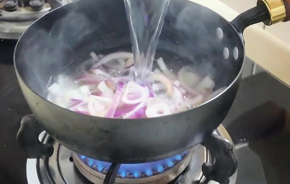 A stream of clear water being poured from a white bowl into a pan containing cooked onions.
